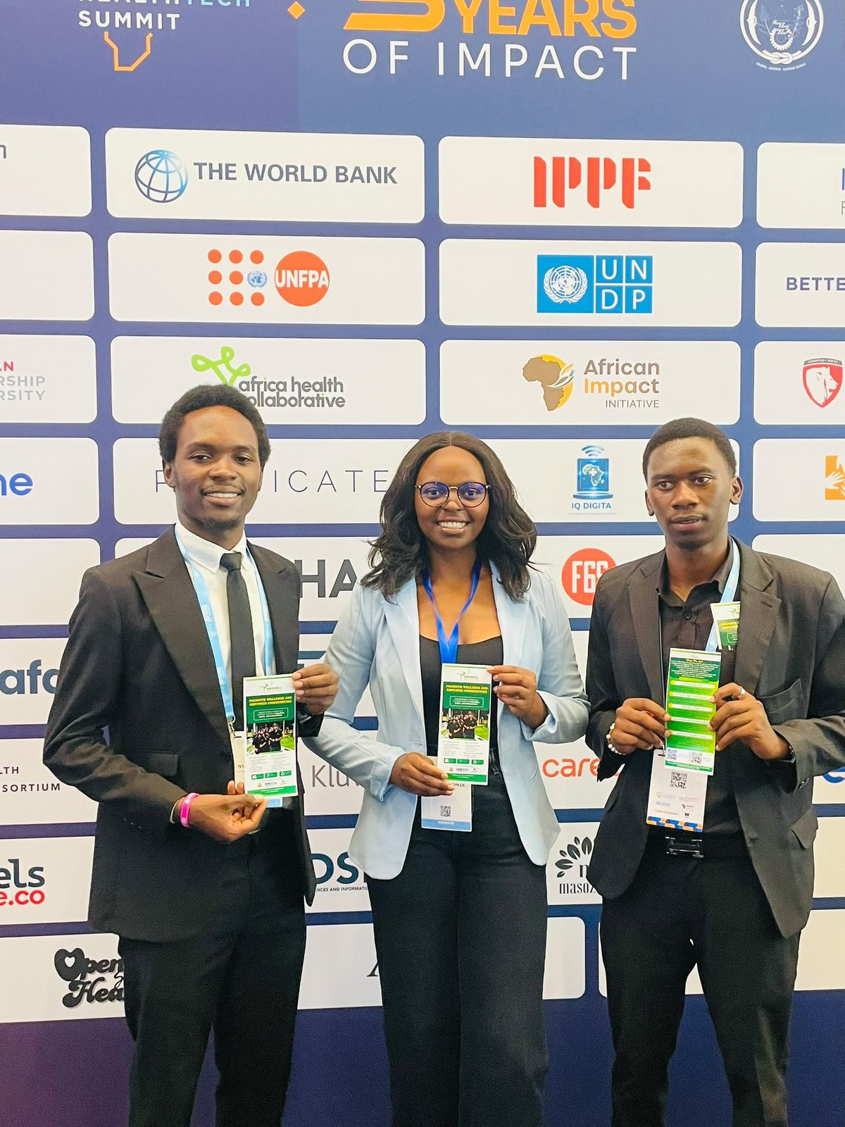 Three MedWell Club representatives in formal attire holding MedWell leaflets in front of the summit backdrop.